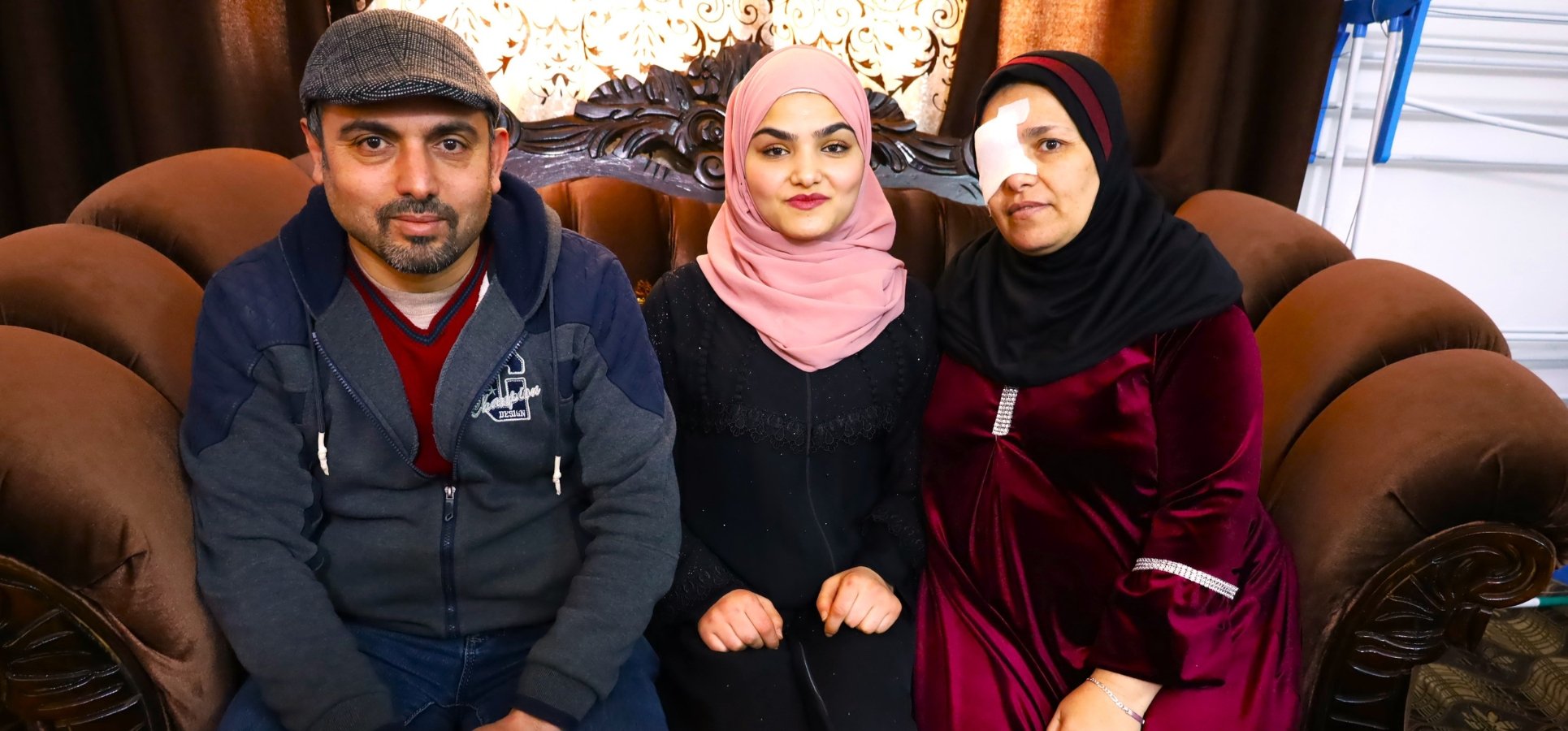 Mariam sitzt zwischen ihren Beiden Eltern auf einem Sofa. Sie hat eine Prothese, ihre Mutter ist an einem Auge verletzt und trägt ein Pflaster.
