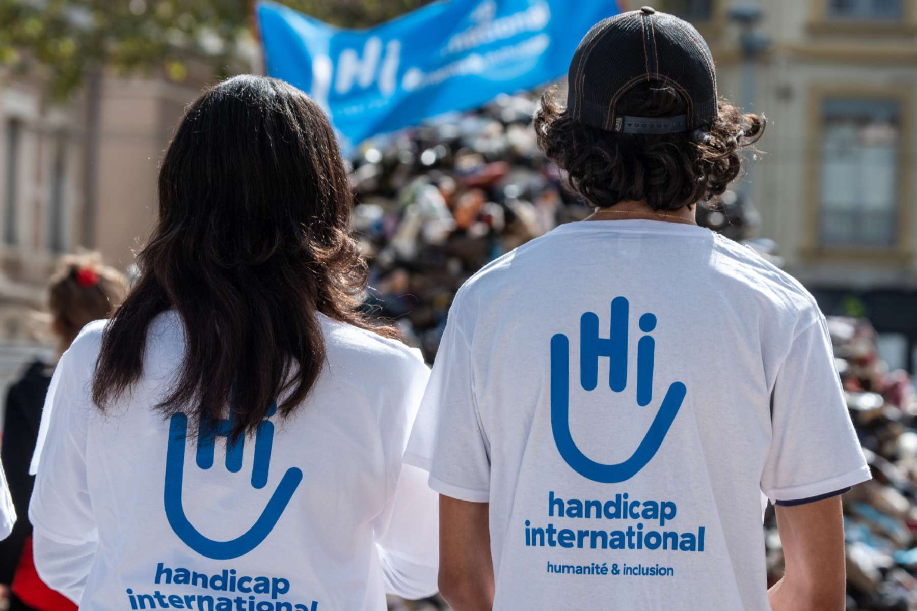 Zwei Hi-Mitarbeiter*innen von hinten. Sie tragen beide das gleiche weiße T-Shirt mit einem großen Handicap-International-Logo auf dem Rücken.