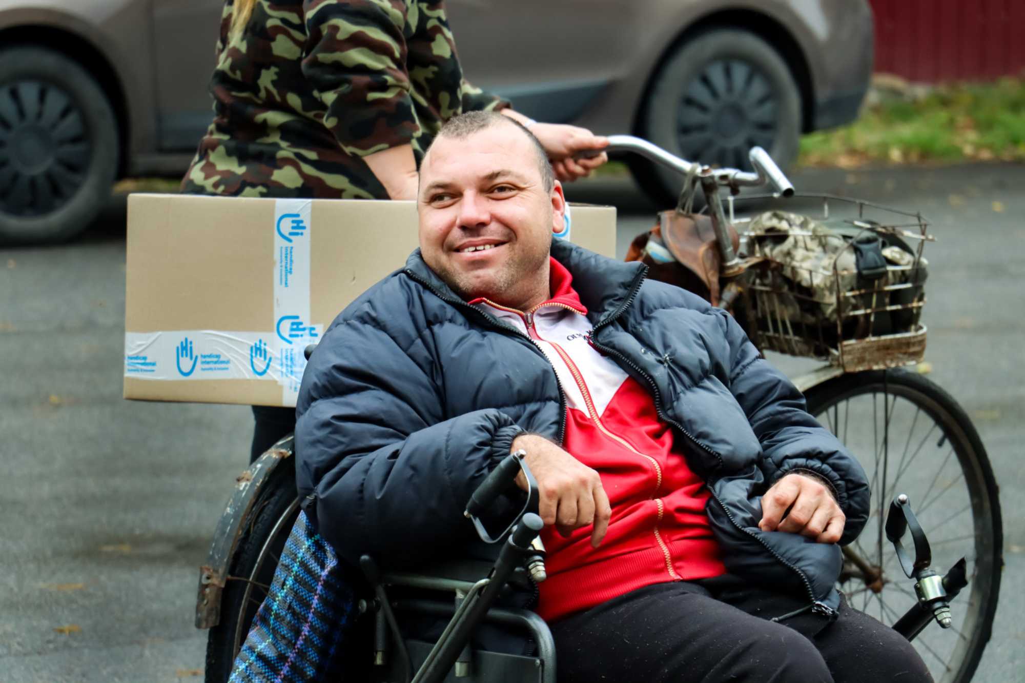 Ein Mann sitzt in seinem Rollstuhl und lacht. Im Hintergrund ist ein Hilfspaket von HI, das auf einem Fahrrad weggeschoben wird.