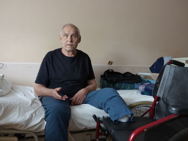 Ein älterer Mann sitzt auf einem Bett. Sein amputiertes Bein hat er auf einem Stuhl daneben abgelegt. 