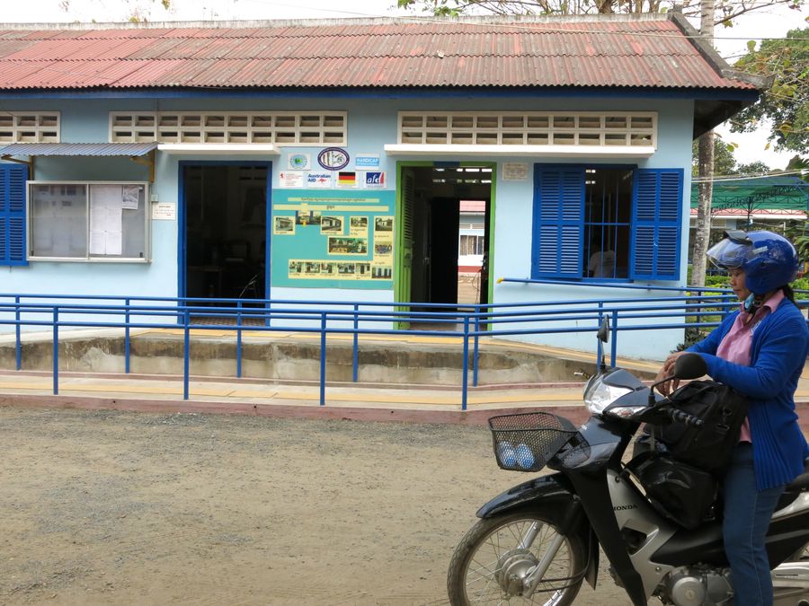 Davann steigt von ihrem blauen Motorroller, im Hintergrund ist ein Rehabilitationszentrum zu sehen. 