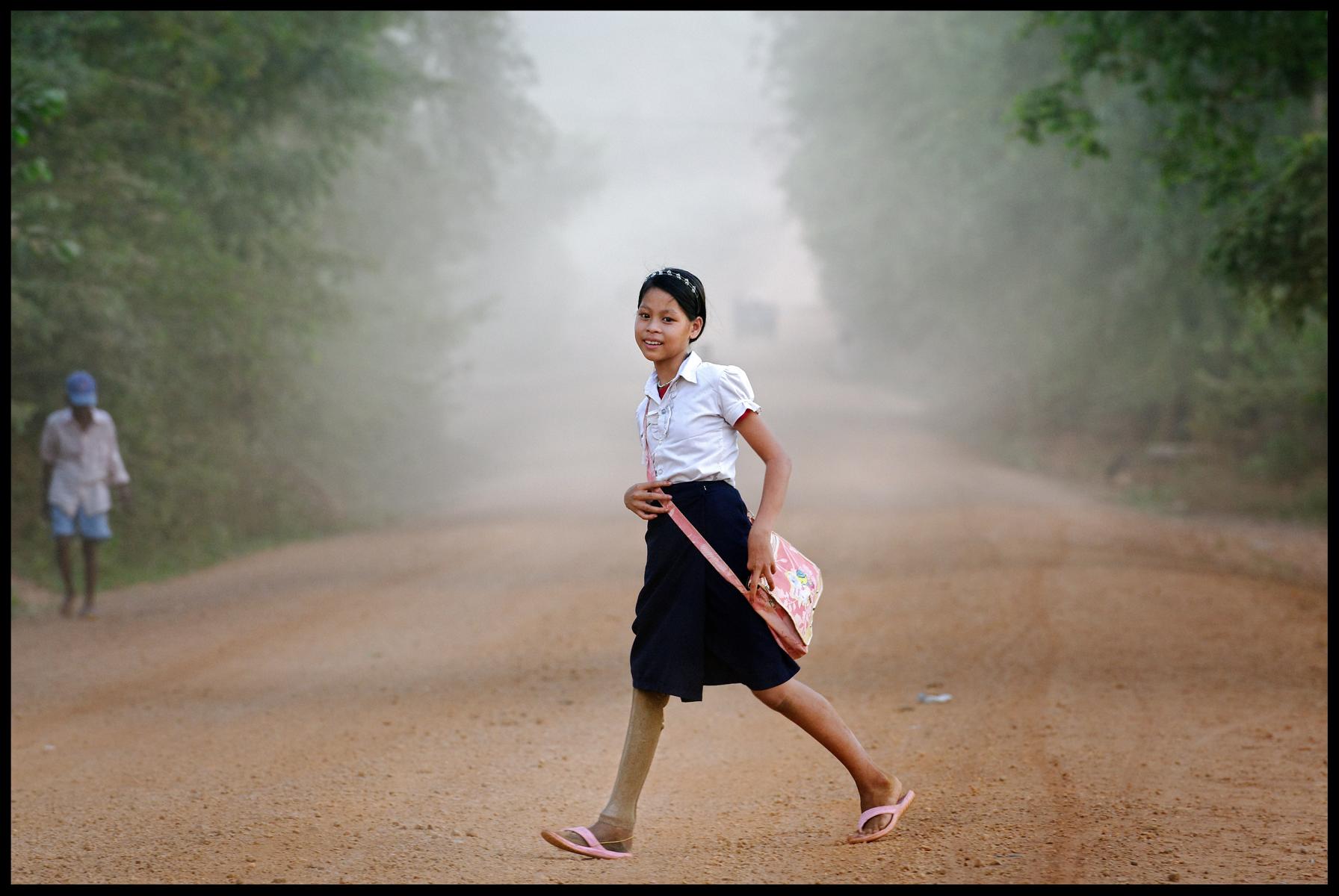 Kanha hat eine Beinprothese. Sie überquert eine Straße, die Schultasche hängt ihr über eine Schulter.