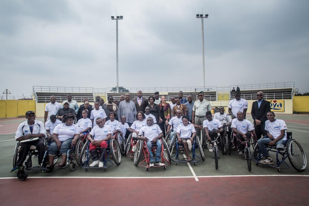 Übergabe der Rennrollstühle. Eine Vielzahl an Athletinnen und Athleten der Paralympics sowie Funktionärinnen und Funktionäre posiert für ein Foto in einem Stadion