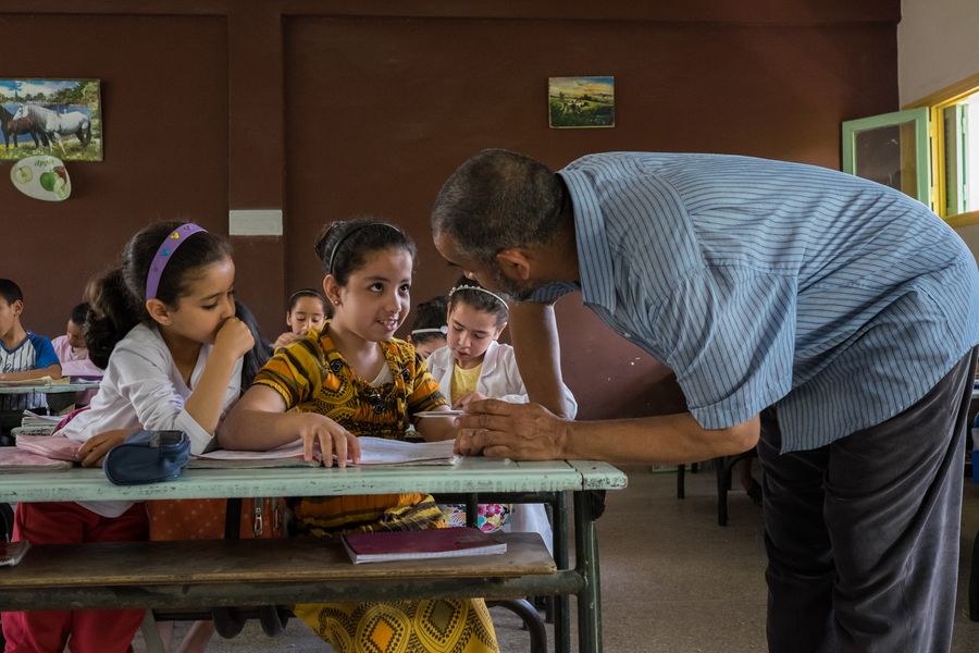 Ikram auf ihrer Schulbank im Unterricht mit ihrem Lehrer. Sie wirkt schüchtern aber konzentriert.