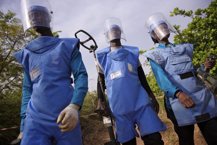 Zu sehen ist Fatou mit 2 Männern. Sie sind in die blaue Sicherheitskleidung für Entminerinnen gkleidet und tragen das Detektoren bei sich. 
