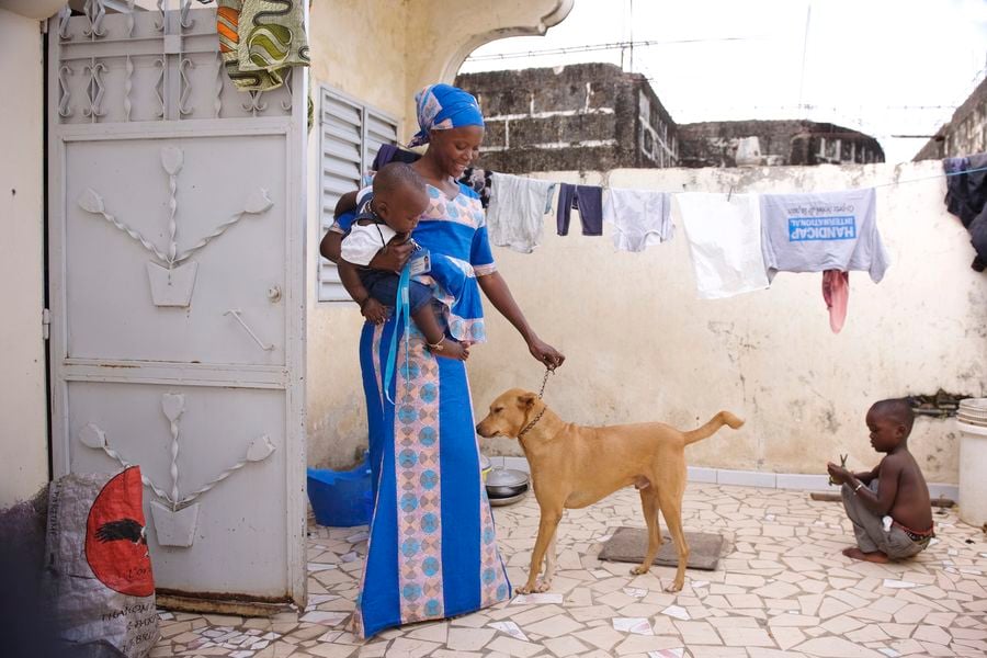 Zu sehen ist Fatou in ihrem Zuhause mit ihrem Hund und ihren zwei Söhnen, einen davon trägt sie auf ihrem Arm. Sie befinden sich auf der Terrasse.