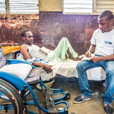 Haiti Matthew inklusive Katastrophenvorsorge Ein Mitarbeitender von Handicap International sitzt auf einem Krankenbett, in dem ein Mann liegt, und notiert sich etwas. Im Vordergrund ist ein Rollstuhl.
