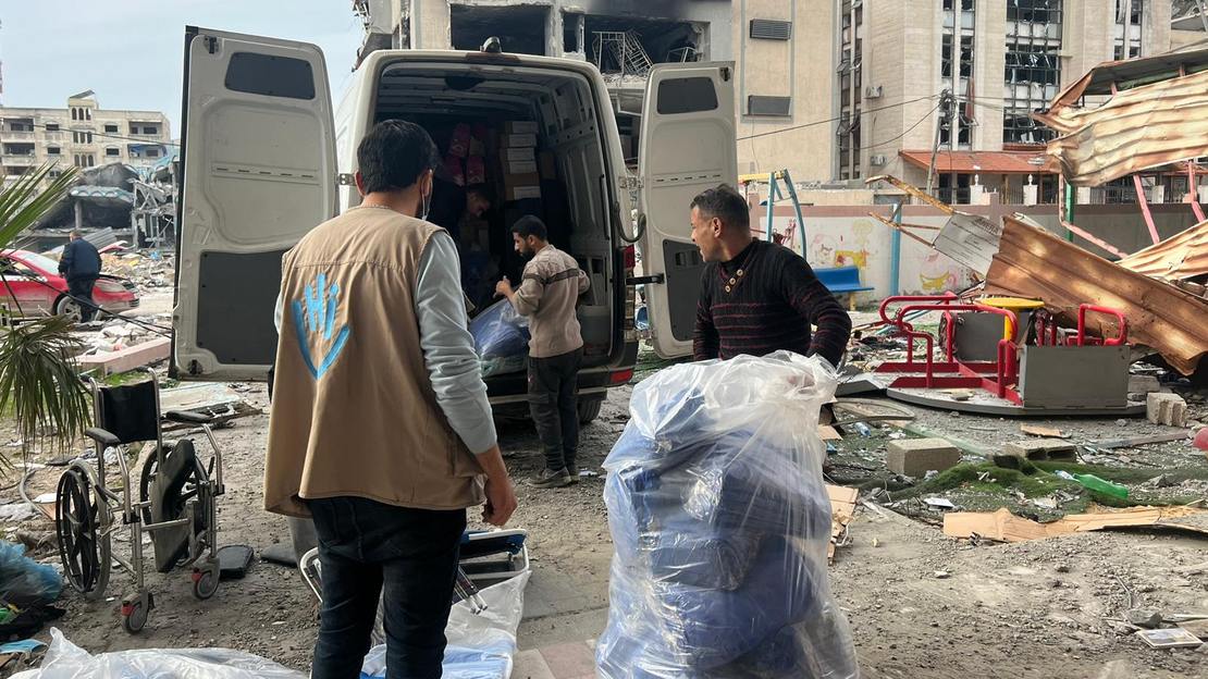Unsere Team laden Rollstühle, Krücken, etc. aus einem Transporter vor einem Krankenhaus. Im Hintegrund liegt die Stadt in Trümmern.