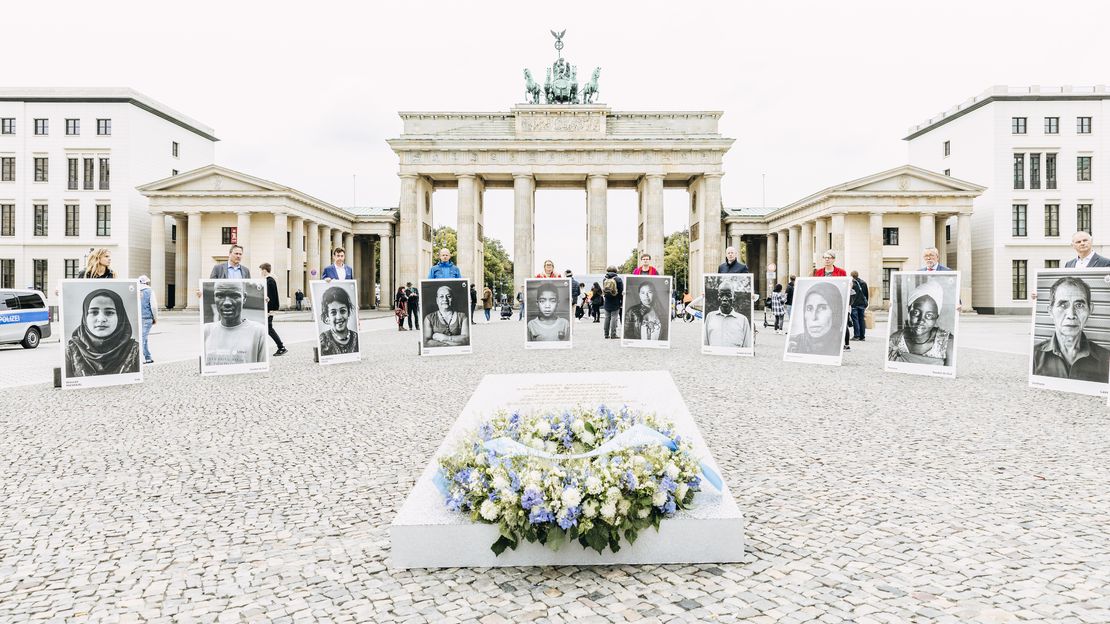 Mahnmal für die vielen unbekannten Opfer von Bombenangriffen auf bewohnte Gebiete. Rund herum stehen Menschen und halten überlebensgroße Porträts von Opfern.