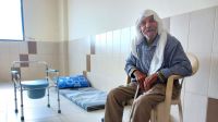 Moustafa Al Sweid, 87 Jahre alt, sitzt auf einem Stuhl und lächelt in die Kamera. Im Hintergrund ist ein Toilettenstuhl.