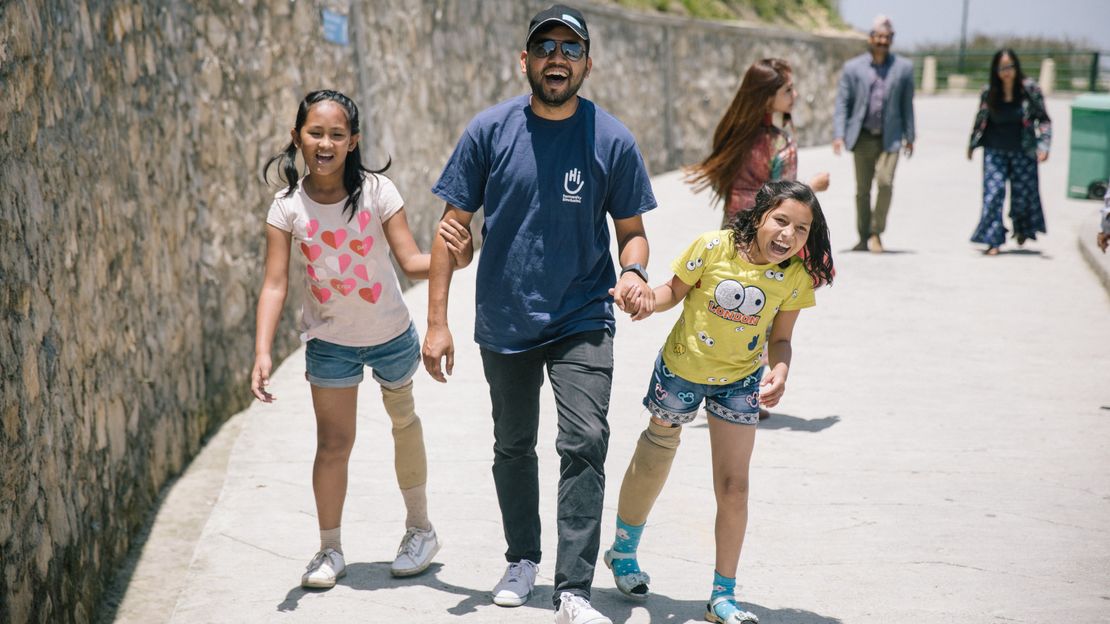 Die beiden Mädchen Nirmala & Khendo aus Nepal gehen Hand in Hand mit ihrem HI-Physiotherapeuten spazieren. HI betreut die Mädchen, seit sie 2016 beim Erdbeben ein Bein verloren haben.