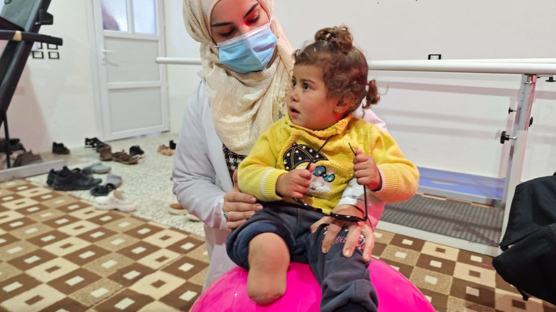 Die kleine Noor sitzt auf einem pinken Gymnastikbild. Ihre Physiotherapeutin ist hinter ihr und hält sie fest.