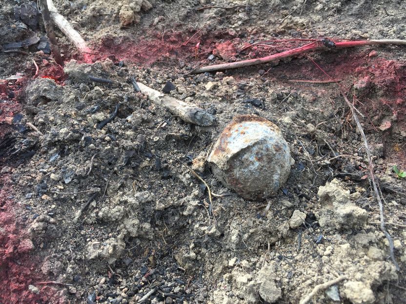 Eine Streubombe liegt schwer erkennbar im Boden zwischen Steinen und Stöcken.