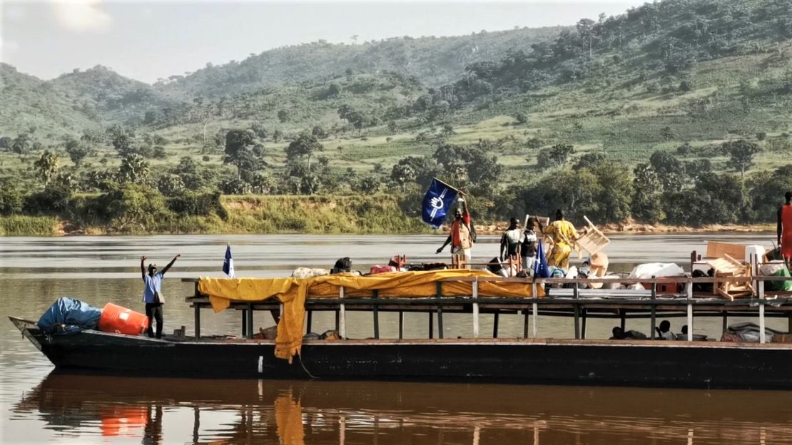 Ein langes Boot auf dem Fluss transportiert Hilfsgüter. Menschen halten eine blaue HI Fahne hoch