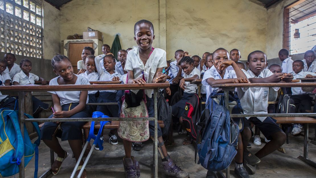 Eine Schulklasse mit vielen Kindern. Im Mittelpunkt steht ein Mädchen mit Orthese am linken Bein. An ihren Tisch lehnen blaue Krücken.