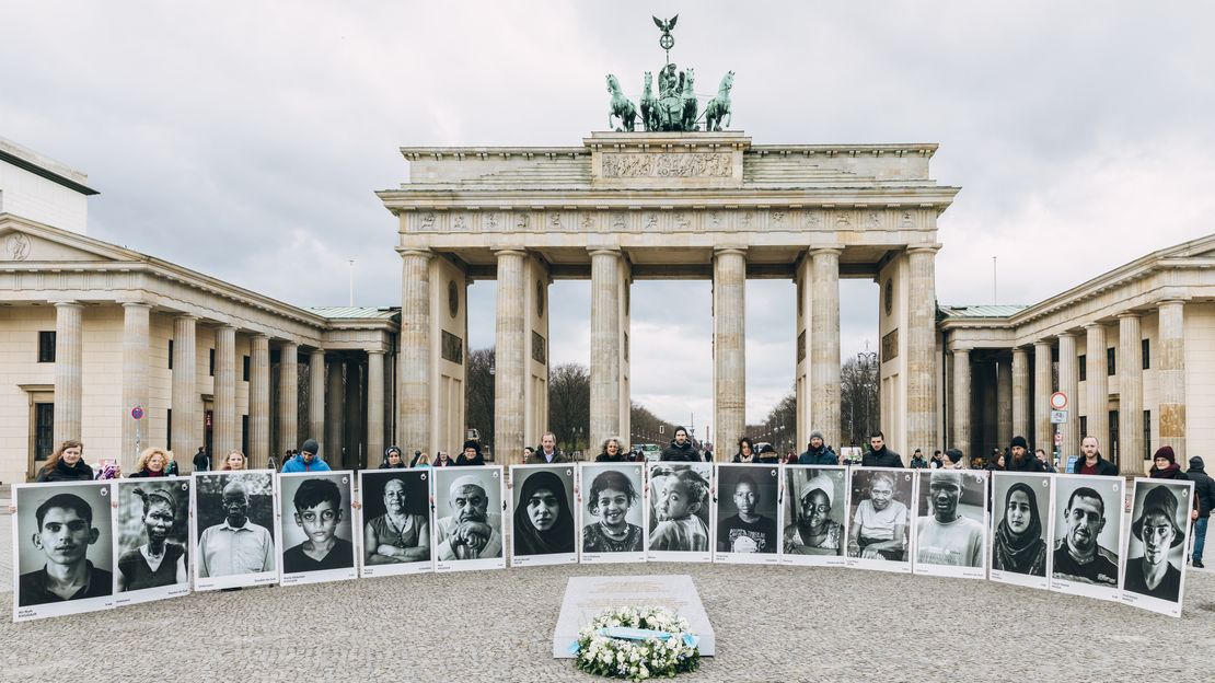 Auf dem Bild sind die Portraits von verletzten und verstorbenen Zivilist*innen aufgereiht vor dem Brandenburger Tor sowie ein Blumenkranz zu sehen.