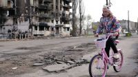 Im Vordergrund ein Mädchen auf einem Fahrrad. Im Hintergrund ist eine zerbombte Häuserfront zu sehen. 