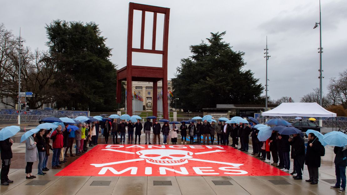 Leute versammeln sich um ein rotes Banner gegen Minen auf dem Boden vor dem Denkmal 'Broken Chair' in Genf vor dem Palais des Nations