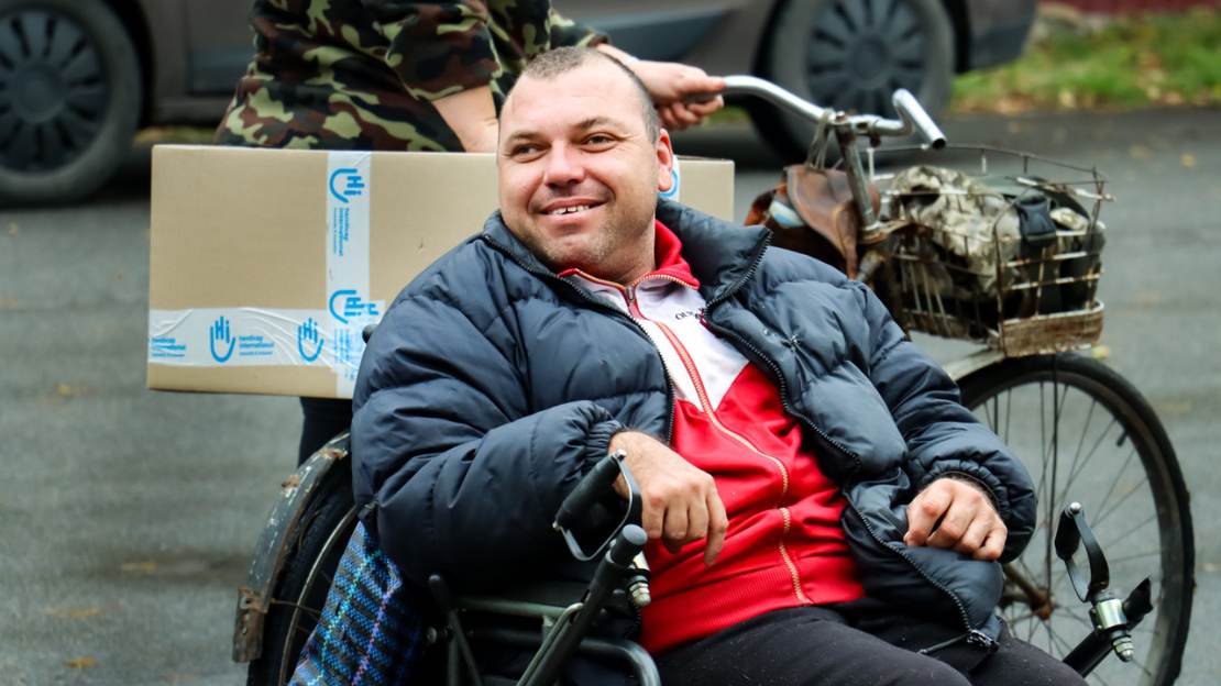 Pavlo sitzt lächelnd in seinem Rollstuhl. Im Hintergrund transportiert jemand sein Paket mit Hygieneartikeln.