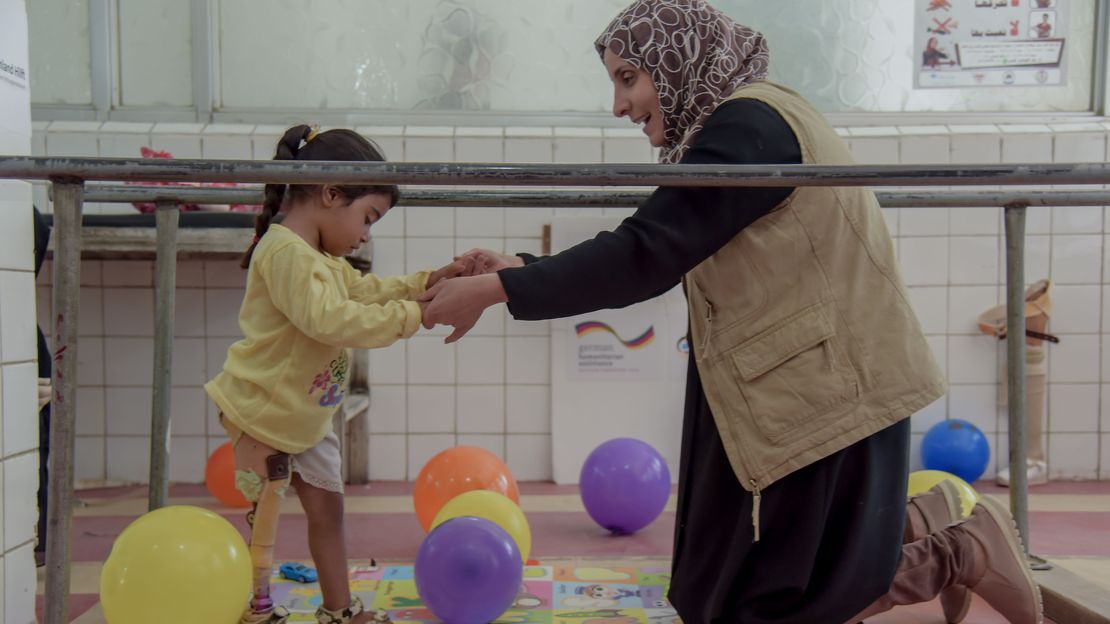 Eine Physiotherapeutin von Handicap International kniet auf dem Boden und hält Halas Hände fest, damit sie das Laufen mit der Prothese üben kann. Auf dem Boden liegen mehrere bunte Luftballons.