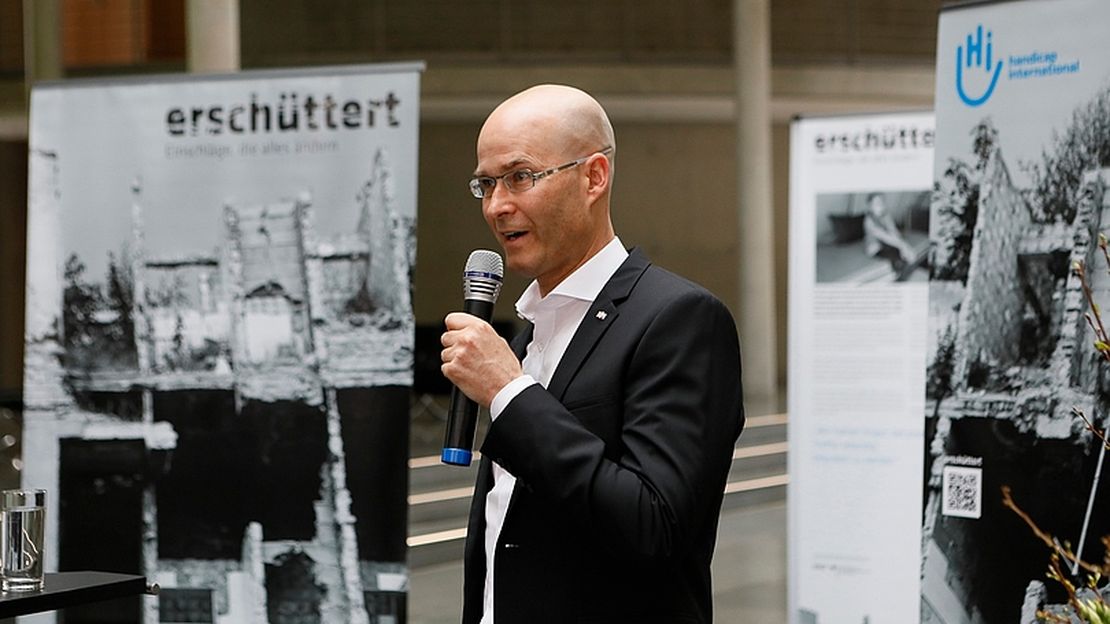 Till Mayer hat ein Mikrofon in der Hand und steht vor den Roll-Ups der erschüttert-Austellung.
