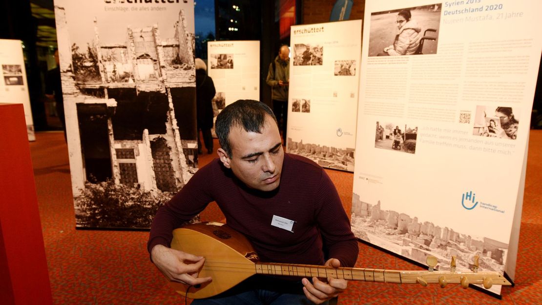 Mohammad sitzt vor den Roll-Ups der erschüttert Ausstellung und spielt auf seinem Instrument. Er hat die Augen geschlossen.