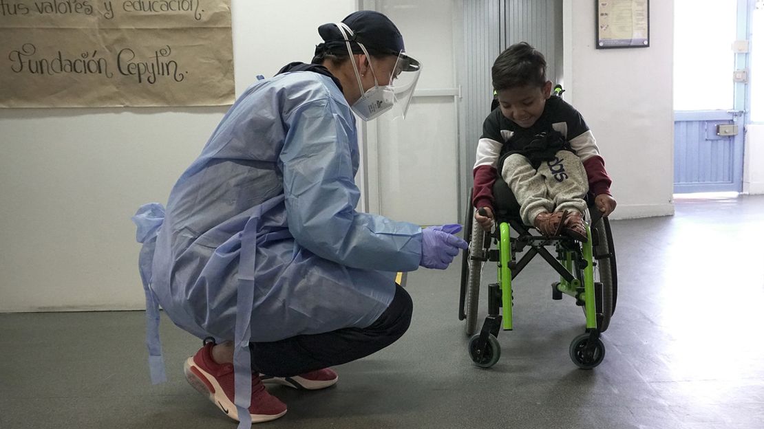 Junge im Rollstuhl, der von einer Physiotherapeutin mit Maske und Schutzkleidung wegen des Coronavirus betreut wird.
