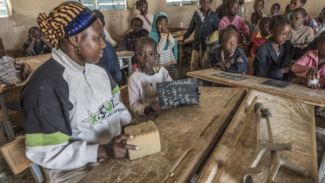 Ein Klassenzimmer mit mehreren Kindern. Eine Lehrerin erklärt ihnen auf einer kleinen Tafel etwas.