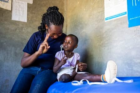 Die kleine Grace in Kenia sitzt auf einer Liege, ihr fehlt ein Bein. Ihre Therapeutin legt einen Arm um sie.