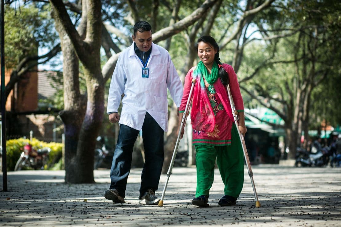 Sanukanchi aus Nepal läuft auf Krücken und mit Prothesen neben einem HI-Physiotherapeuten. Sie lächelt. Sie hat nach der Geburt keine Behandlung ihres Klumpfußes erhalten.