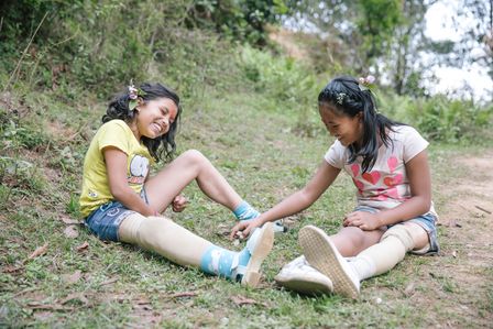 Nirmala und Khendo aus Nepal tragen beide eine Beinprothese. Sie sitzen lachend auf einer Wiese.