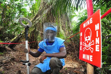 Ein HI-Entminer sitzt in voller Ausrüstung vor einem Schild, dass vor Blindgängern warnt.
