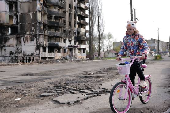 Ein Mädchen auf einem Fahrrad. Im Hintergrund ein zerbombtes Wohnhaus.