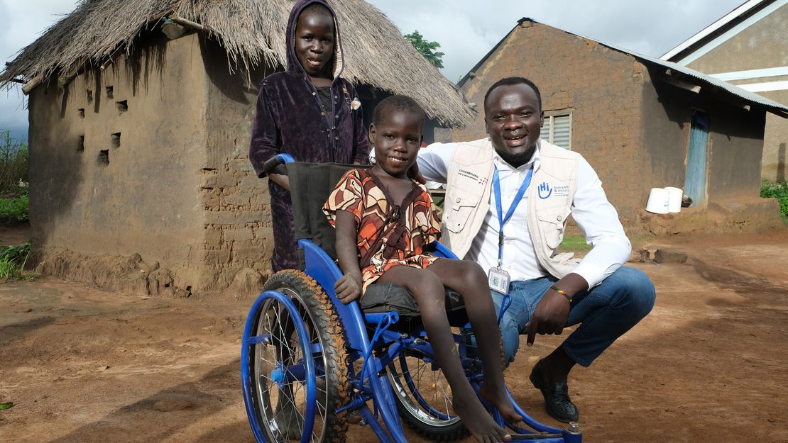 Ein Mädchen sitzt in einem Rollstuhl. Hinter ihr steht ihr Schwester und neben ihr ein Physiotherapeut von HI.