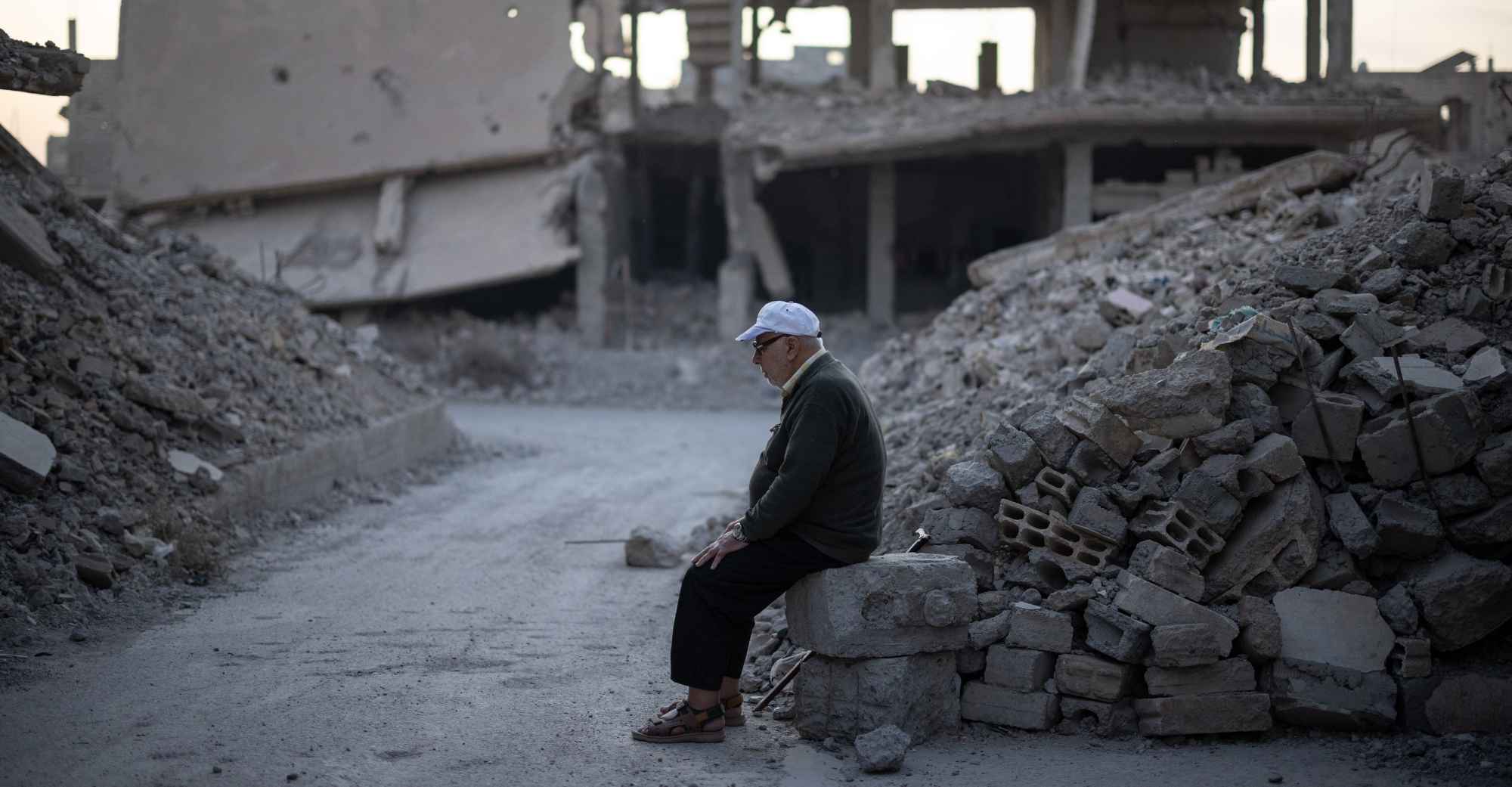 Ein älterer Mann sitzt auf einem kleinen Trümmerhaufen. Um ihn rum stehen Häuserruinen. 