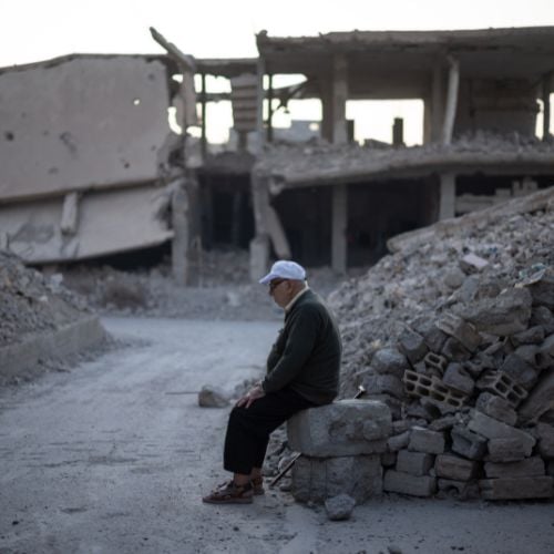 Ein älterer Mann sitzt auf einem kleinen Trümmerhaufen. Um ihn rum stehen Häuserruinen. 