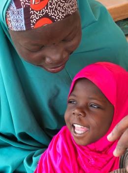 Massalouka in knallpinkem Kopftuch sitzt lachend auf dem Schoß ihrer Mutter Massoura