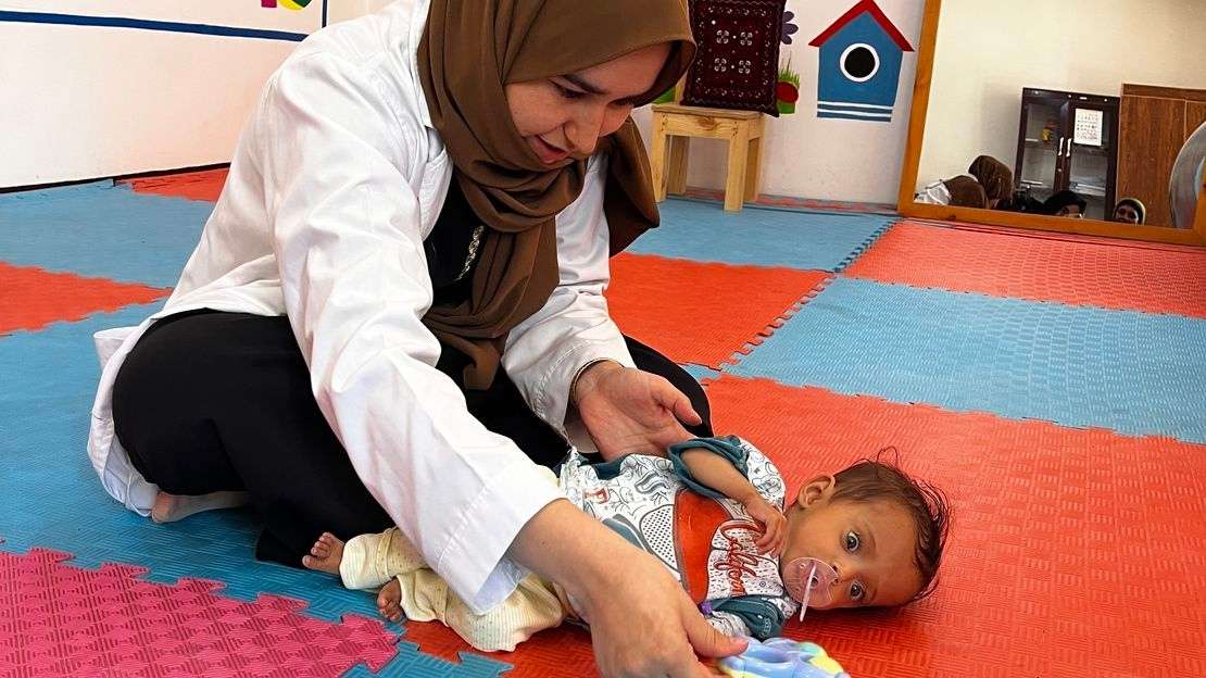 Ein Baby liegt auf einem gepolsterten Boden in einem Rehazentrum. Über dem Baby lehnt eine Frau und hält ihm ein Spielzeug hin. 