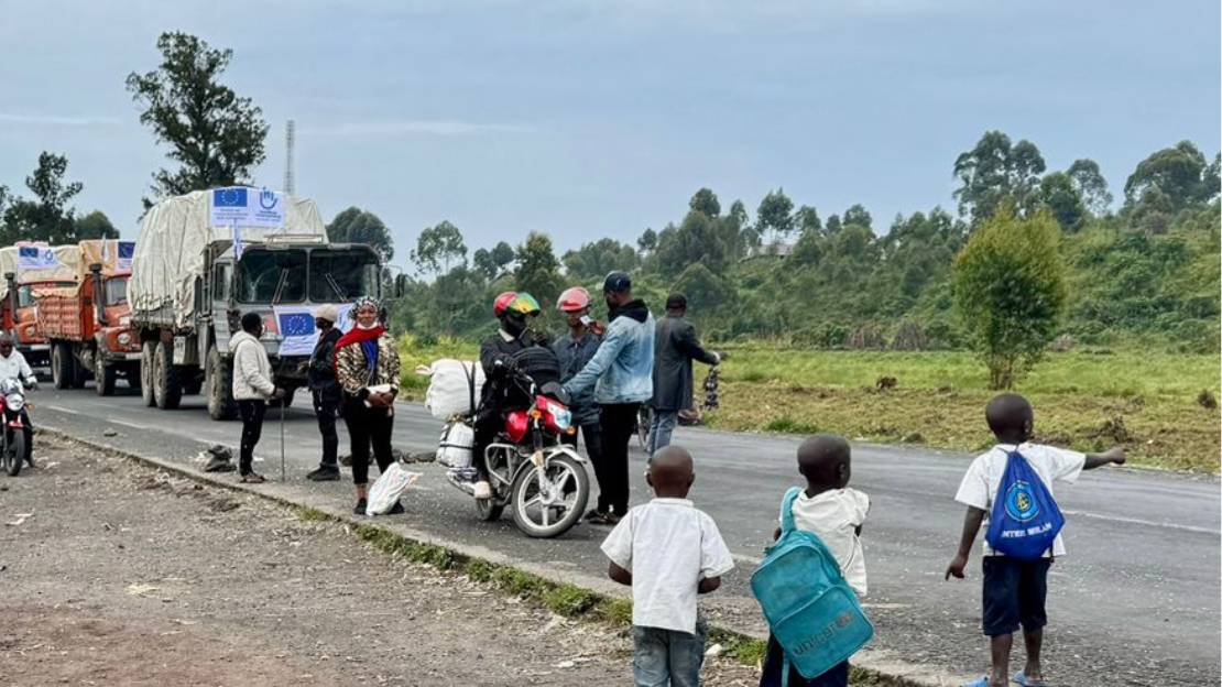 Beladene LKWs mit Logos von Handicap International fahren eine Straße entlang. Ringsherum stehen viele Menschen. 
