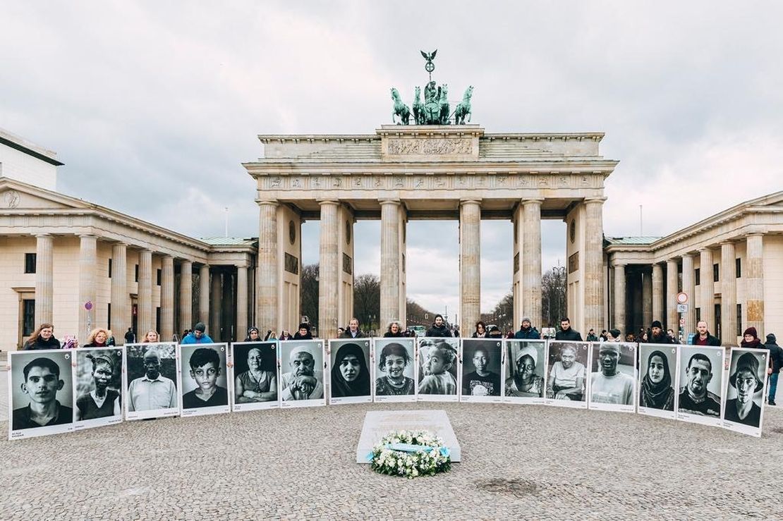 Mitarbeiter/-innen von Handicap International halten überlebensgroße Opferporträts hoch und haben sich um das Mahnmal für die unbekannten Zivilist/-innen vor dem Brandenburger Tor aufgestellt
