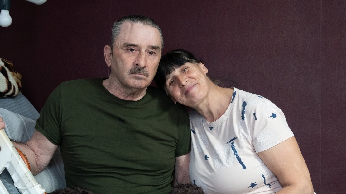 Mann und Frau sitzen auf einem Krankenbett. Die Frau lehnt sich lächelnd an ihren Mann.