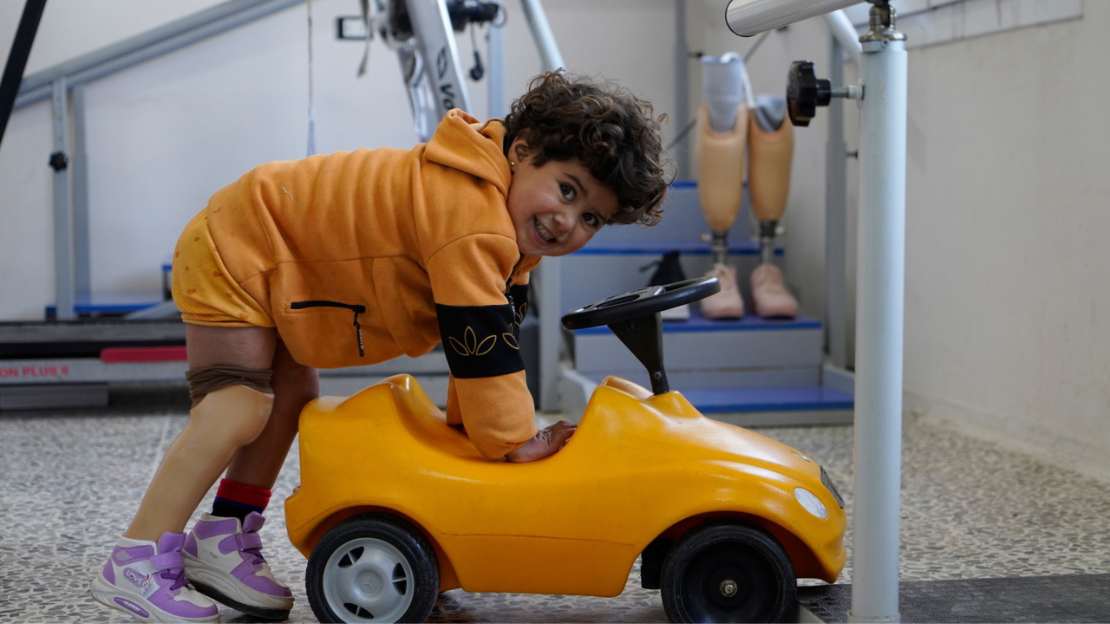 Ein kleines Mädchen mit dunklen Locken und einer Beinprothese lehnt auf einem Spielauto und lacht. 