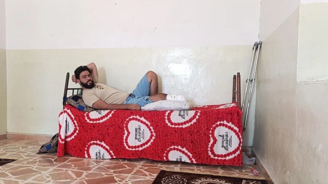 Ein Mann liegt auf einem einfachen Bett. Sein eines amputiert und verbunden. Er schaut ernst. 