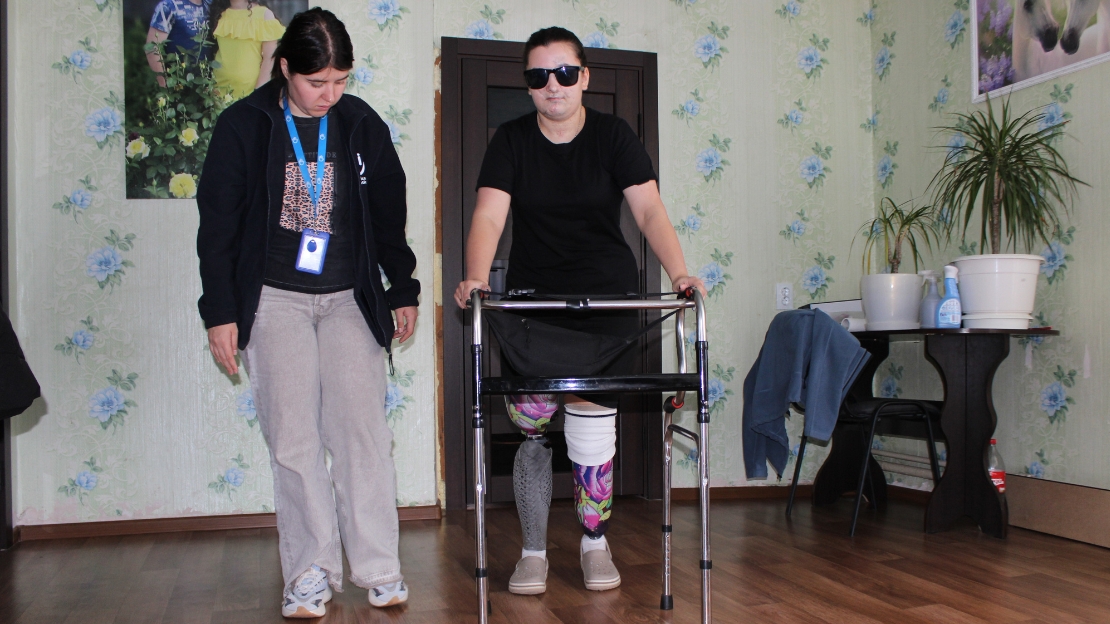 Yuliia, eine junge Frau mit Sonnenbrille, nutzt einen Rollator und trägt jeweils eine Prothese an beiden Bein, während eine HI-Physiotherapeutin daneben steht und sie unterstützt.