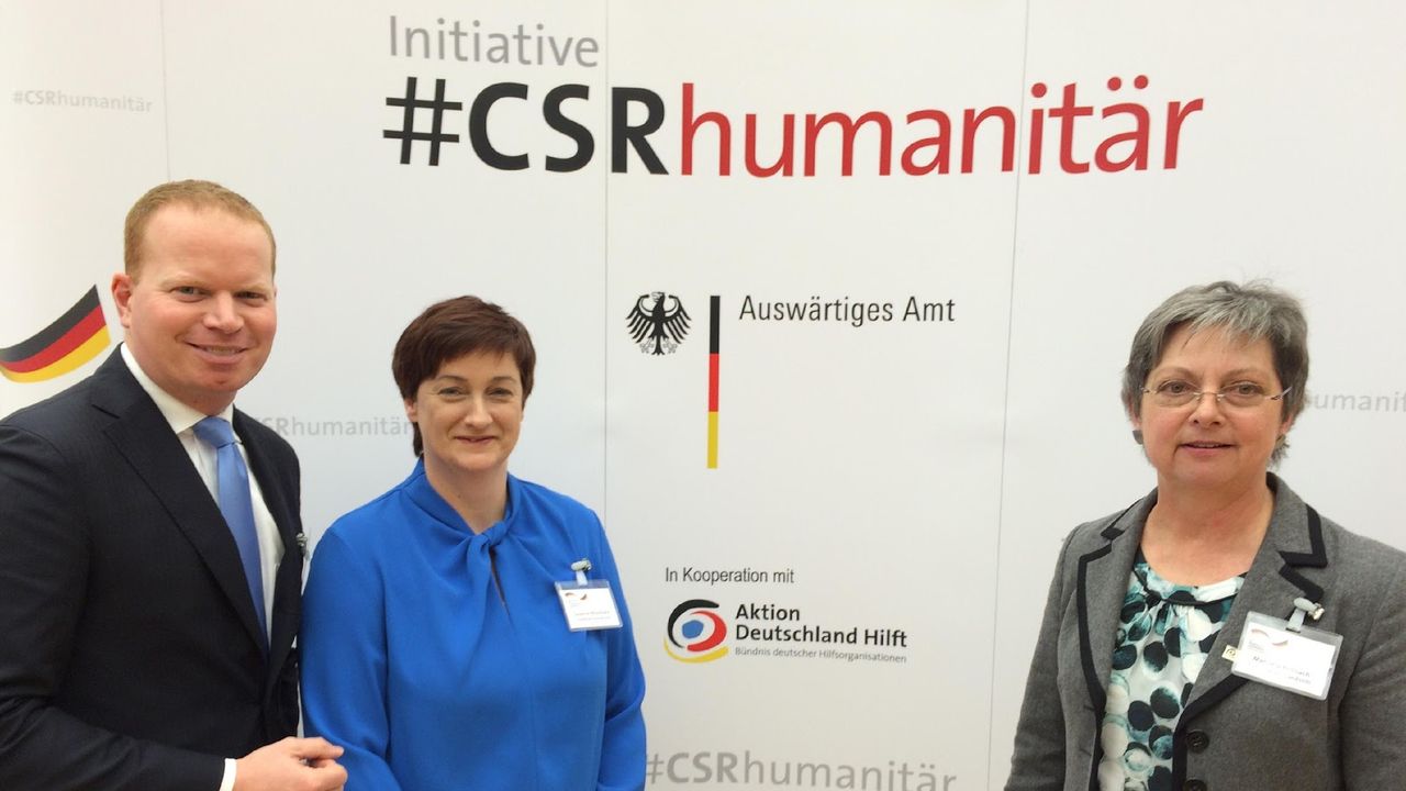 Ein Mann und zwei Frauen stehen vor einer Werbetafel des CSR-Forums. Der Mann steht im Anzug, neben einer Frau, die eine blaue Bluse trägt, ganz links im Bild. In der Mitte das CSRhumanitär Logo. Rechts im Bild steht eine etwas ältere Frau, die in grau gekleidet ist.