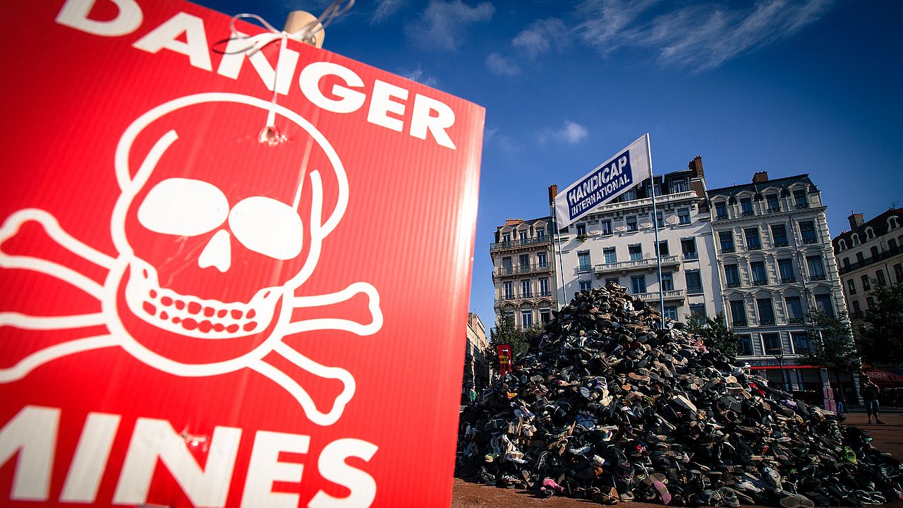 Zu sehen ist ein Danger Mines Zeichen mit einer Protestschuhpyramide im Hintergrund und einem Handicap International Emblem auf der Spitze.