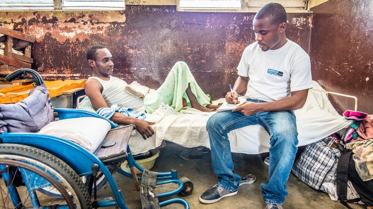 Ein Mitarbeitender von Handicap International sitzt auf einem Krankenbett, in dem ein Mann liegt, und notiert sich etwas. Im Vordergrund ist ein Rollstuhl.