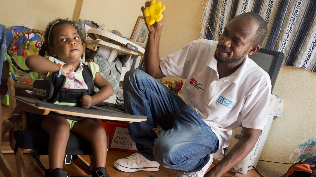 Rehabilitationstechniker Franz besucht die kleine Patientin Mella in Port-au-Prince. Er kniet neben ihr und spielt mit Mella.