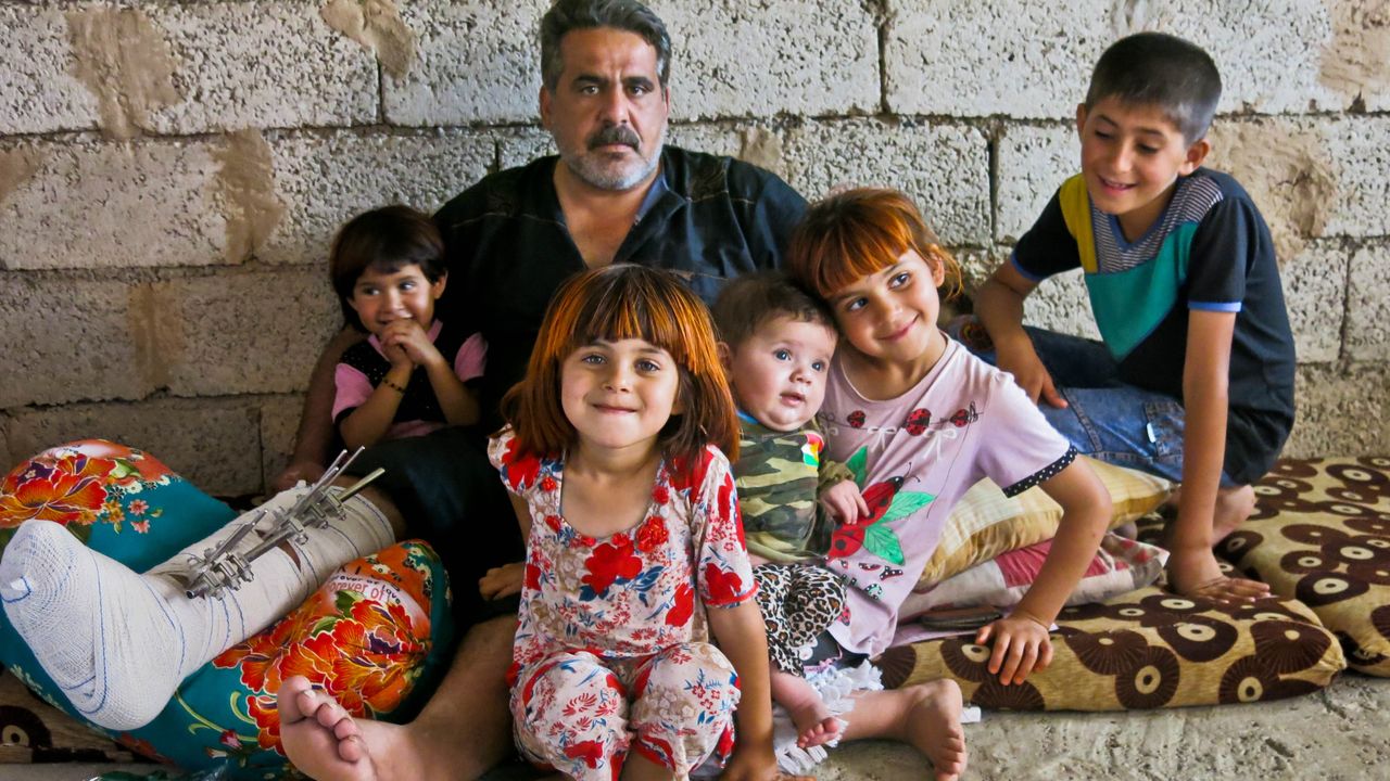Saadi sitzt mit seinen Kindern auf dem Boden. Er umarmt sie väterlich. Sie wirken glücklich.