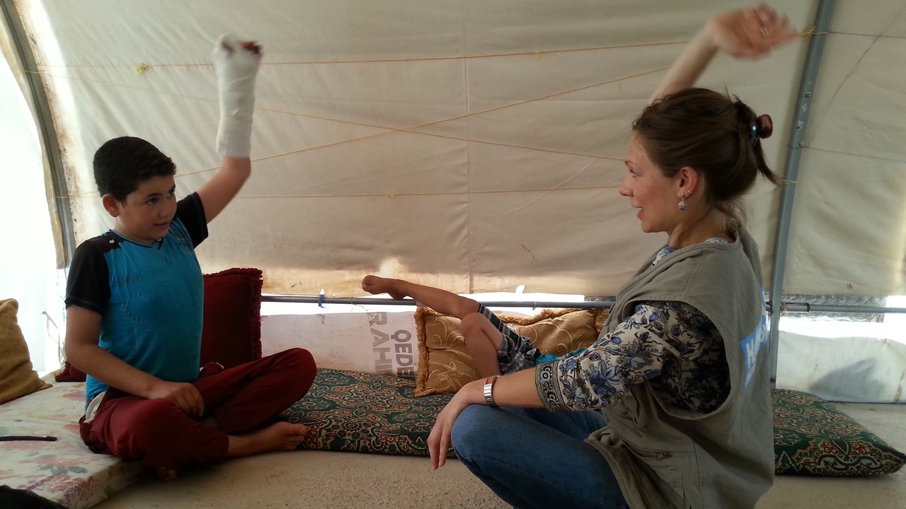 Saddam sitzt seiner Physiotherapeuting von Handicap International im Schneidersitz gegenüber. Beide strecken einen Arm in die Luft. Saddams linker Arm ist einbandagiert.